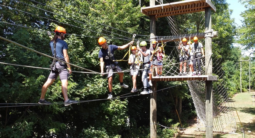 A group of people wearing safety gear are attached by ropes on the obstacle of a high ropes course. 